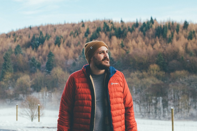 Man with a moustache and beard wearing a toque and a puffy jacket with snow and trees in the background