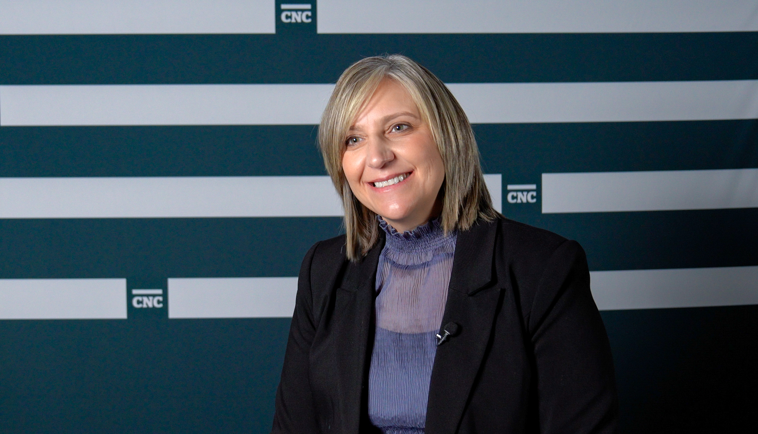 President Cindy Heitman sits smiling in front of a teal backdrop with white stripes and CNC logos